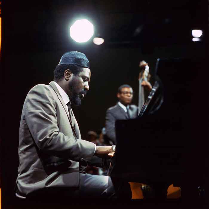 Thelonious Monk sitting at a piano wearing sunglasses and a marroon hat