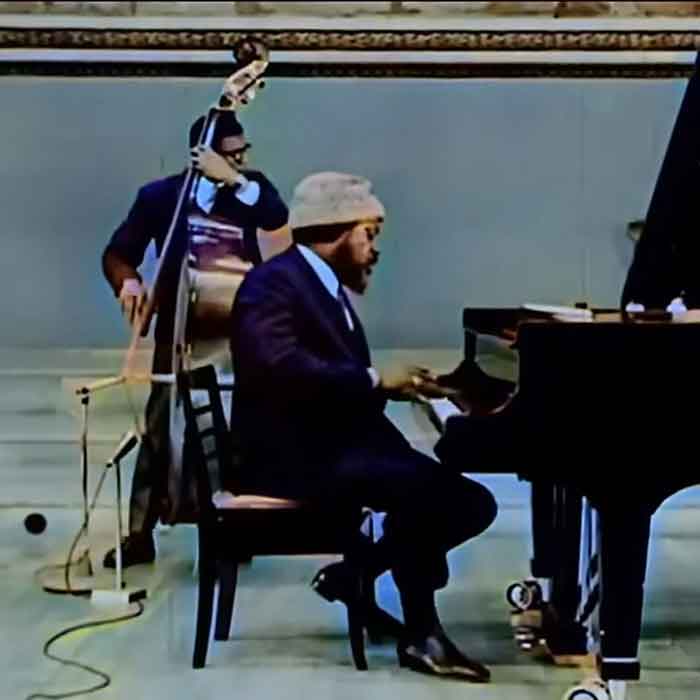 A side view of Theolonious Monk playing the piano with an upright bass player on a stage