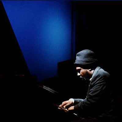 Thelonious Monk playing piano on a stage with a blue background. he is wearing a black hat.