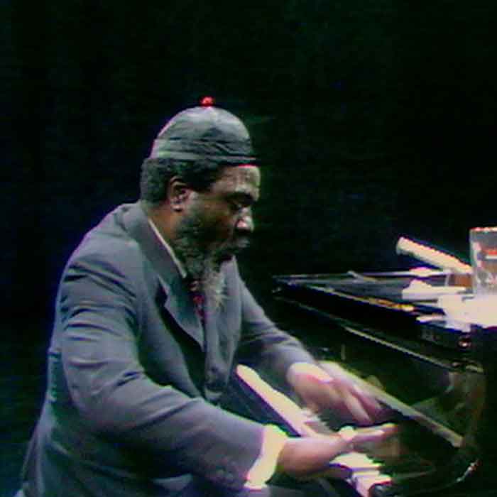 A side view of Theolonious Monk playing the piano with an upright bass player on a stage